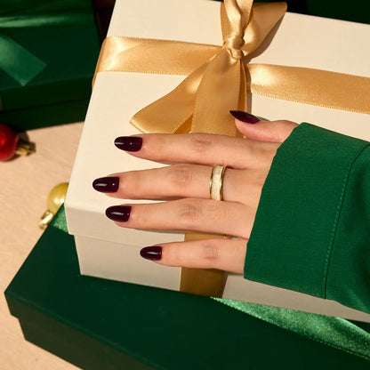 Hand with dark nail polish holding a gift box with a gold ribbon, wearing a gold ring.
